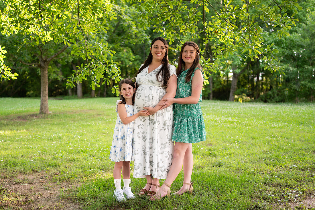 Expecting mother standing with two girls beneath leafy trees